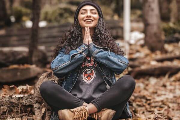 Donna sorridente seduta in un bosco con le mani in namastè: lo yoga ci aiuta a vincere lo stress e a cominciare a vivere bene