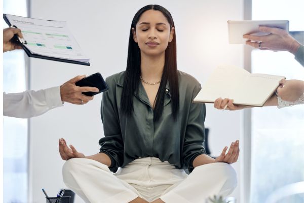 Donna che medita seduta sulla scrivania mentre attorno tutti le porgono qualcosa da fare: respiro yoga nella vita quotidiana